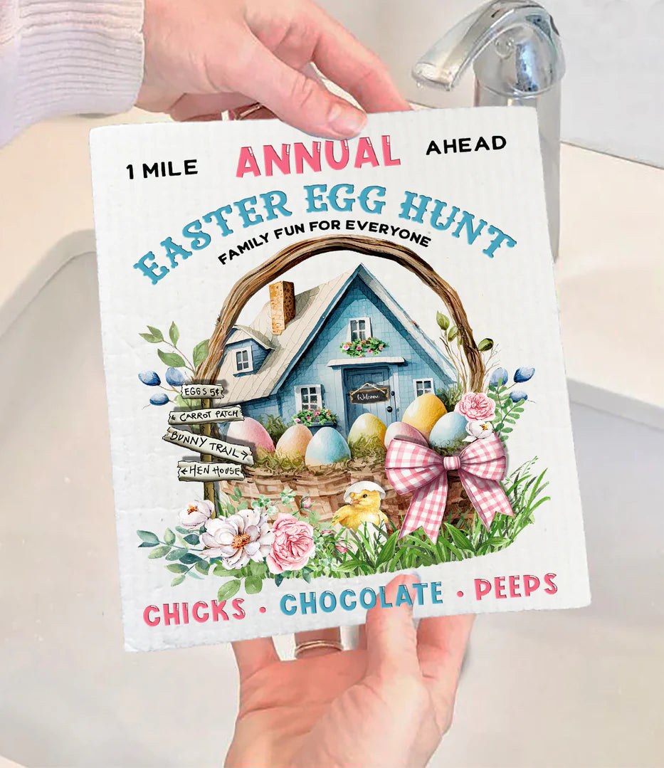 A person holding a Swedish dishcloth with an annual Easter egg hunt theme, featuring a blue farmhouse, chicks, eggs, and the text 'Chocolate + Peps'.