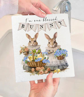 A white Swedish dishcloth with a floral design and two bunny figures, held by a person. The text on the dishcloth says 'I'm blessed to have bunny's as neighbors' surrounded by a floral wreath.