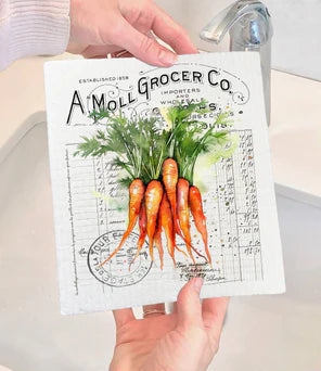 A person holding a white Swedish dishcloth with a vintage farmhouse design featuring carrots and other vegetables.
