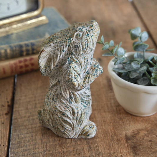 A figurine of a praying bunny made from concrete, displayed on a wooden surface with a plant pot and book in the background.