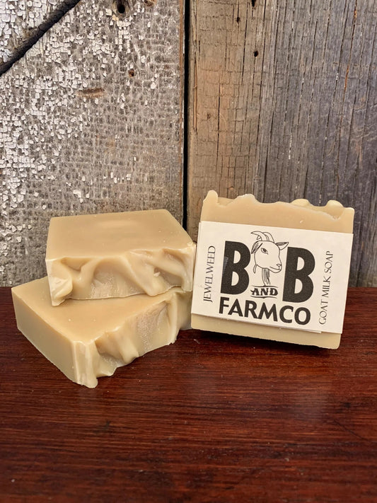Two bars of soap with the B&B Farmco brand logo on the packaging, placed on a wooden surface with a rustic background.