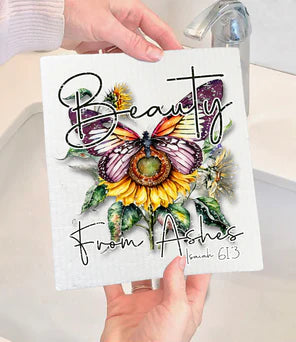 A person holding a white Swedish dishcloth with a colorful butterfly design and the text 'Beauty from Ashes' along with a verse.