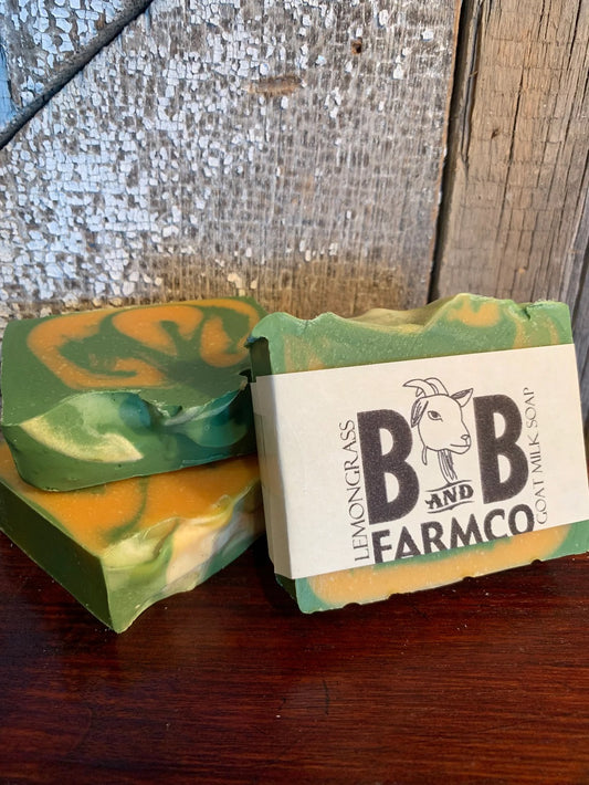 A bar of goat milk soap with a lemongrass scent, featuring a green and yellow swirled design, placed on a wooden surface with a branded label on top.